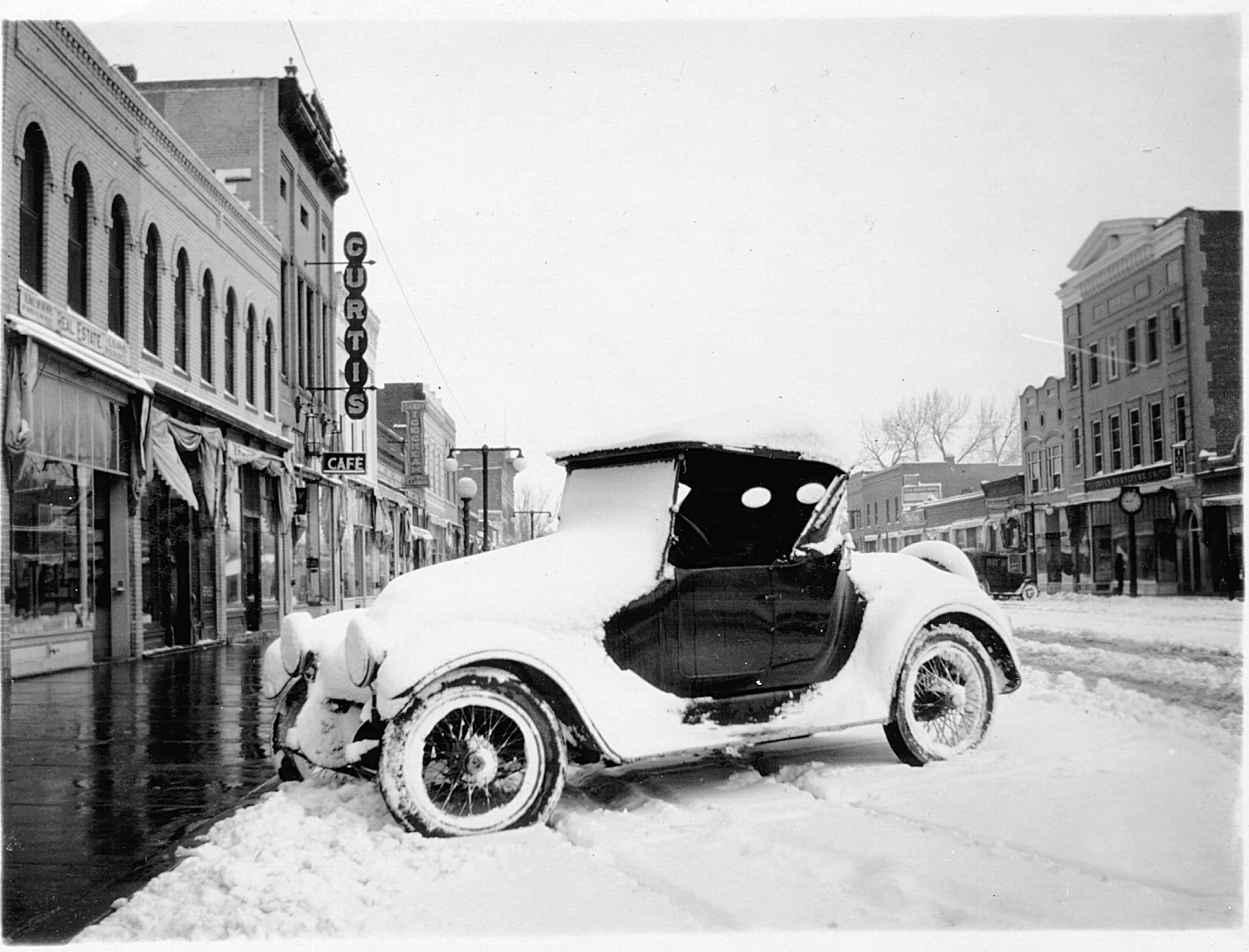 Historic black and white photo of a 1920s car on Main Street in the snow