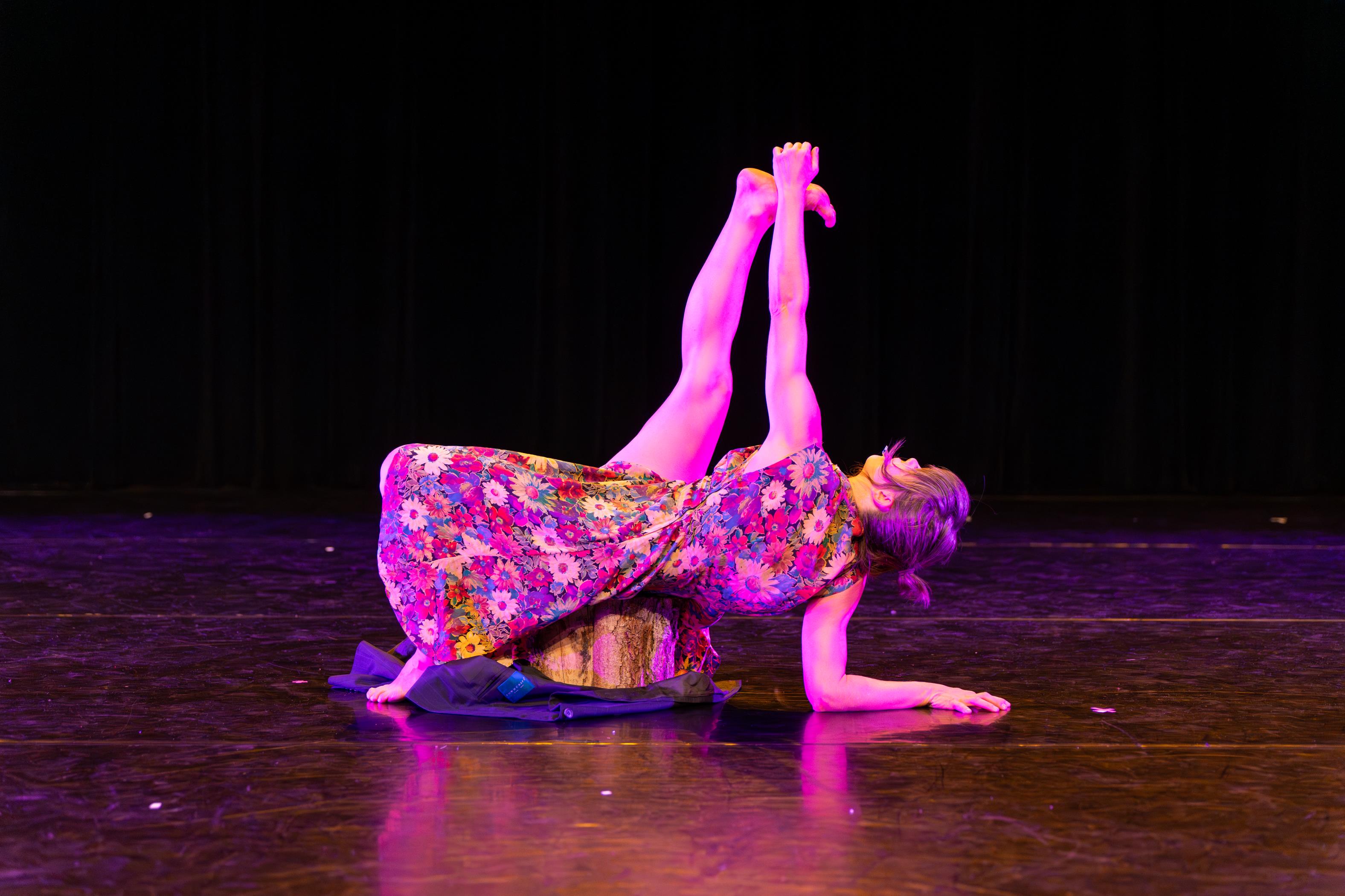 Mariposa Collective dancer on stage stretching her arm to her extended leg in a pose atop a tree stump.