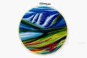 Colorful felted Maroon Bells landscape with colorful foreground of green, blue, red, yellow, green, and white and a mountainous background of green, blue against a blue an white sky.