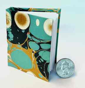 Miniature book bound in a colorful cover with gold, turquoise, black, and white orb design, Book shown next to a quarter to illustrate scale.