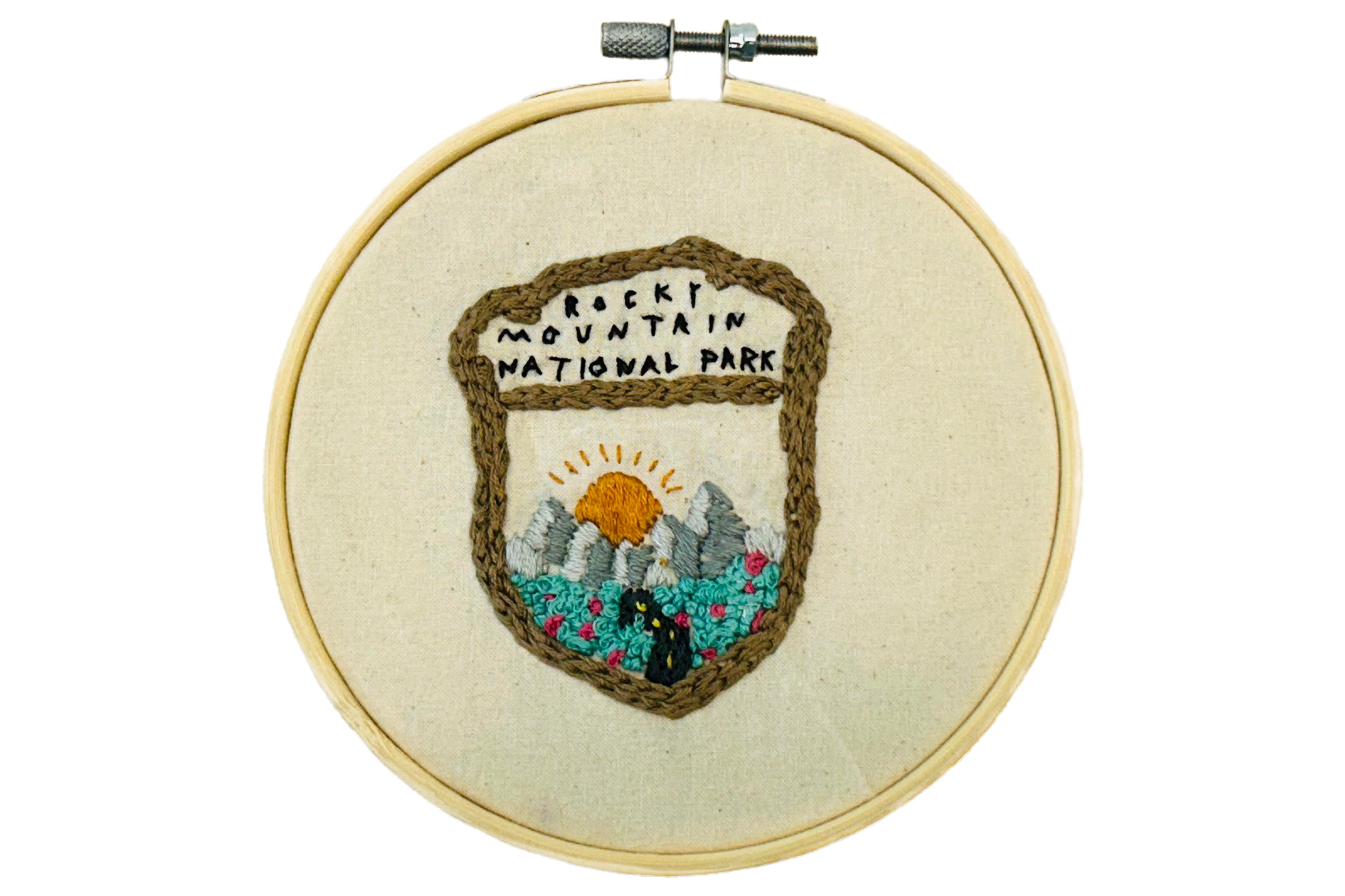 Embroidery displayed in hoop of Rocky Mountain National Park badge featuring a winding road surrounded by lush grass and pink flowers leading to the mountains with the sun peaking out behind them.