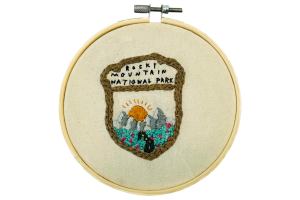 Embroidery displayed in hoop of Rocky Mountain National Park badge featuring a winding road surrounded by lush grass and pink flowers leading to the mountains with the sun peaking out behind them.