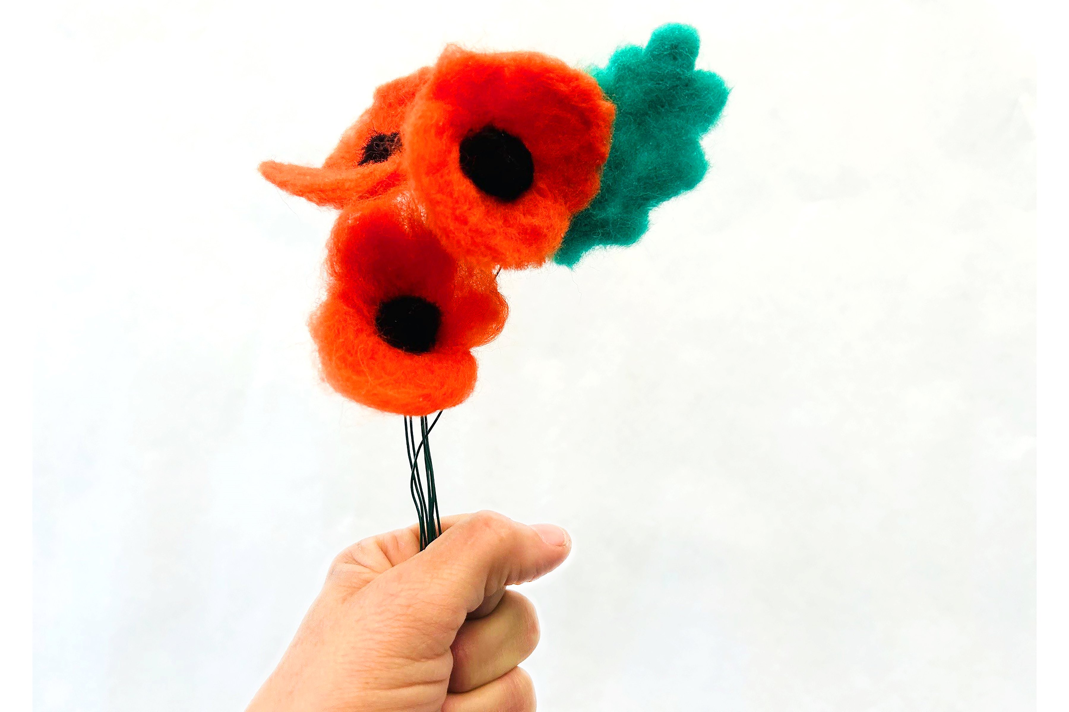 Needle felted arrangement containing three poppies (red with black center) and a green leaf attached to wire stems.