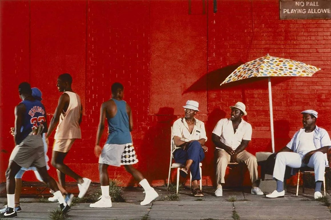 A group of four young men pass by a group of three older men sitting in lawn chairs shaded by an umbrella.