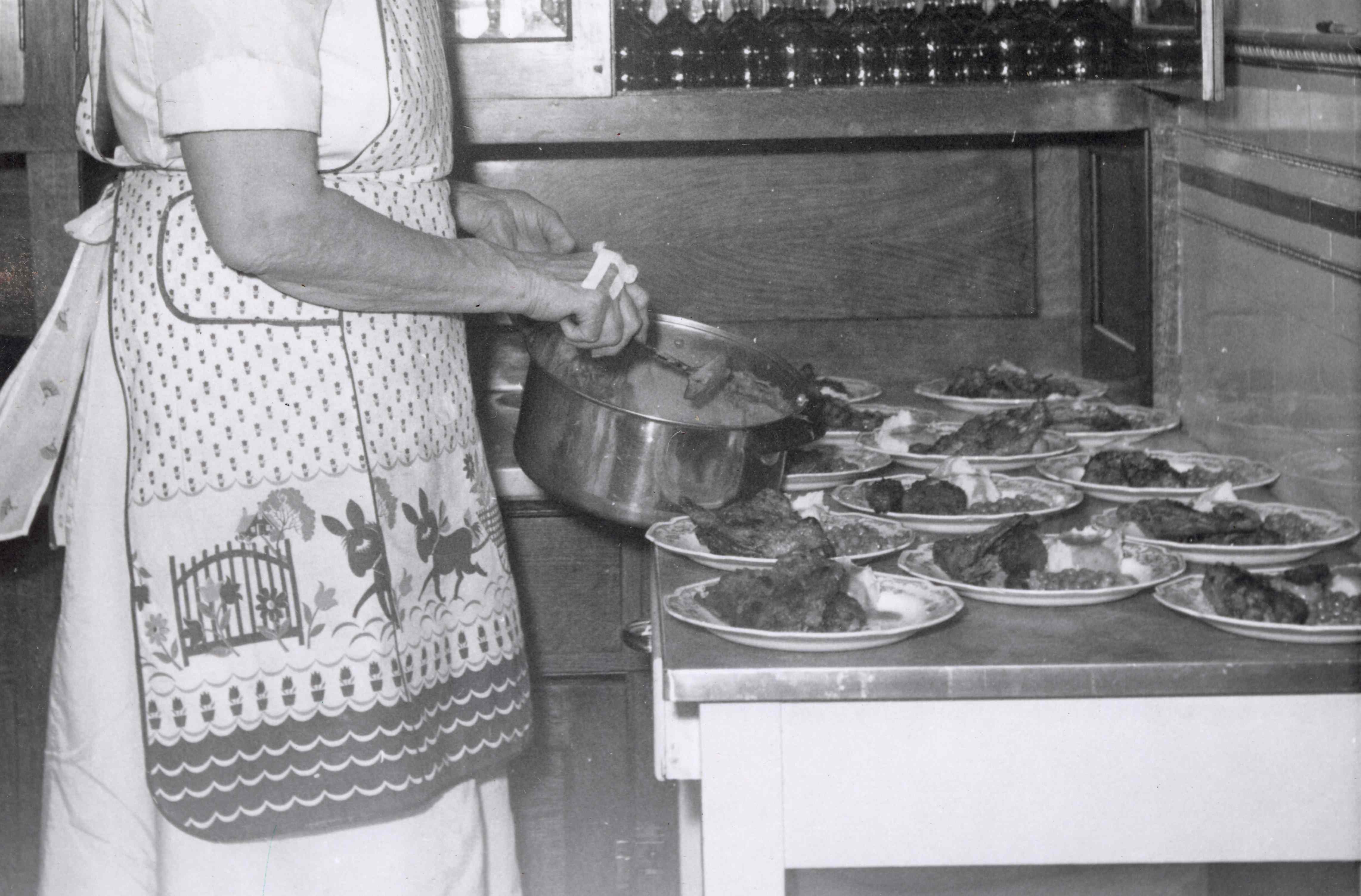 A historic black and white image of a woman wearing an apron backing pies