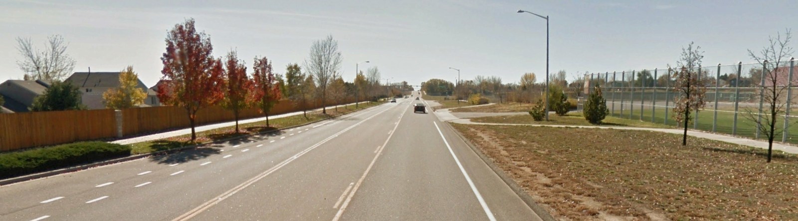 A street with trees and grass on each side of the road. To the left is a fence with houses behind and the right side of the road shows a field behind a chain link fence.