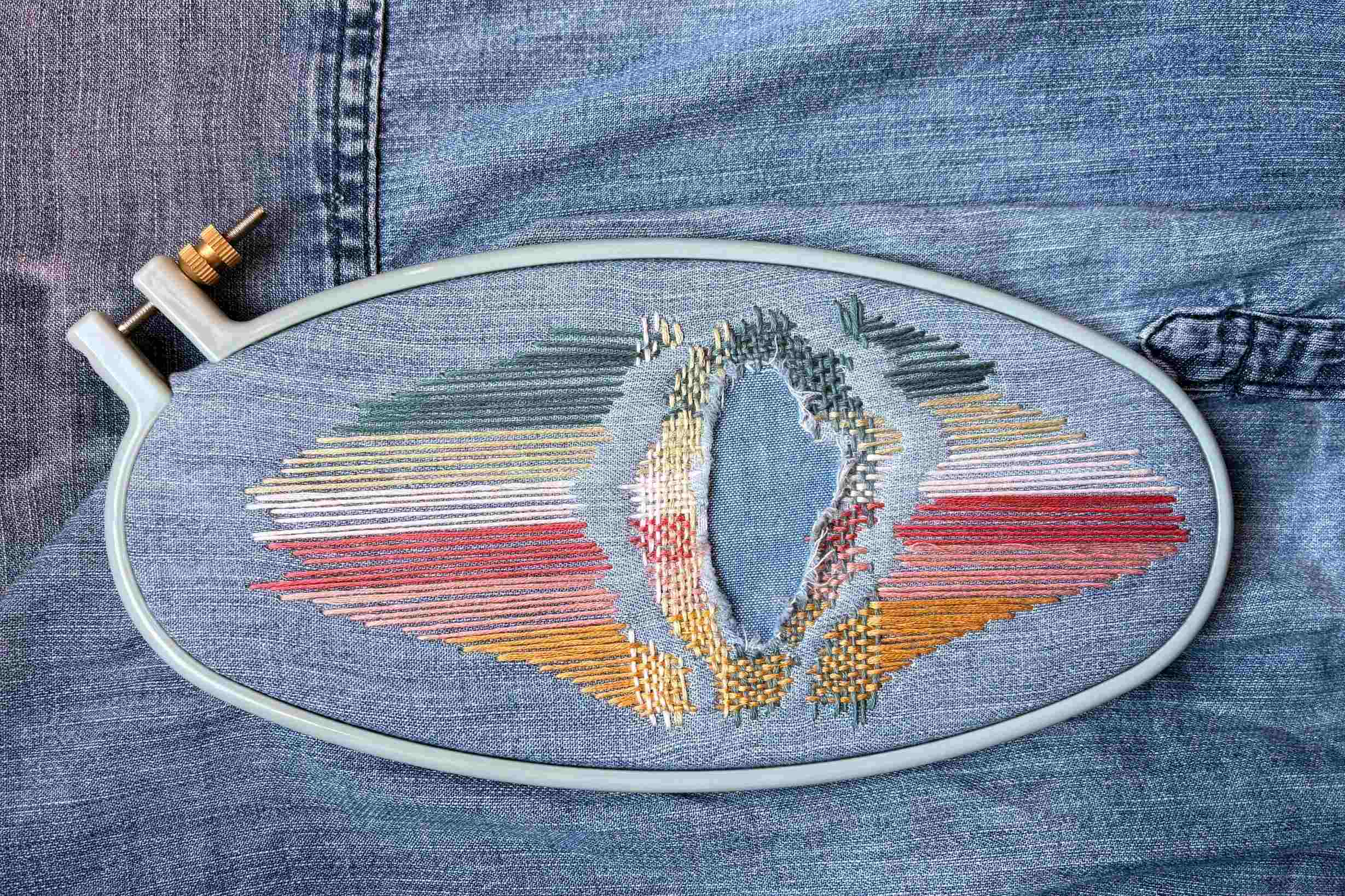 Rainbow stitched artistic repairs of a tear in a piece of denim. Stitching framed by oval embroidery hoop.