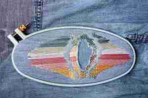 Rainbow stitched artistic repairs of a tear in a piece of denim. Stitching framed by oval embroidery hoop.