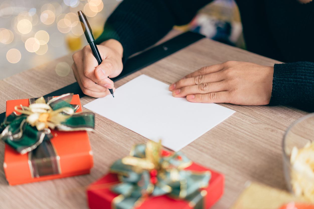 A pair of hands begin to write a letter with a pen. The paper sits on a table that includes two wrapped presents, while the background has out-of-focus golden decorations in the background.