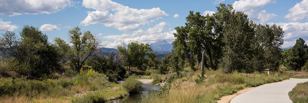 A paved trail runs alongside a wooded creek with mountains in the background.