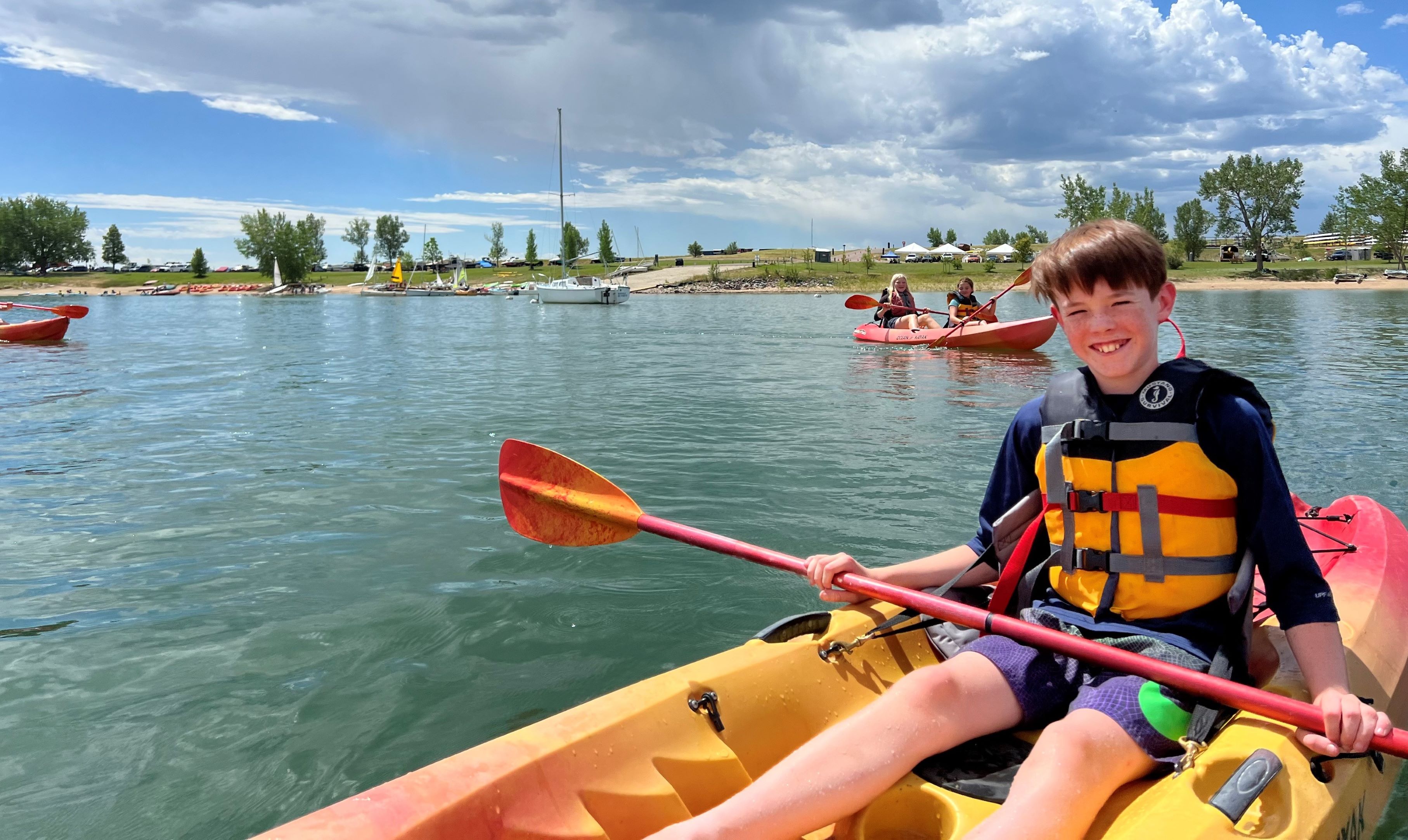 Young boy in a life jacket, in a kayak on a lake looking at the camera. Other kayakers in the background.