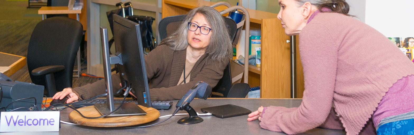 Library visitor getting help at the Help Desk at the Library next to a welcome sign.