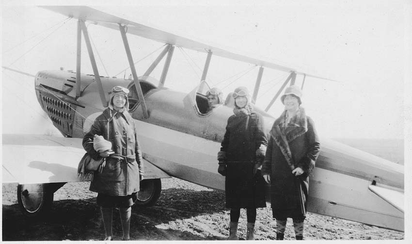 Black and white snapshot of three women standing beside a biplane with the male pilot in the cockpit. The woman on the left is wearing a canvas aviator's cap with goggles perched on top of her head.