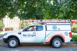 NextLight truck on street beneath a tree, with the driver waving through the window.