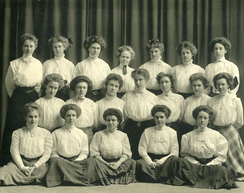 Photograph of 19 young women in 1908 wearing white blouses and dark skirts standing, kneeling, and sitting in three rows.