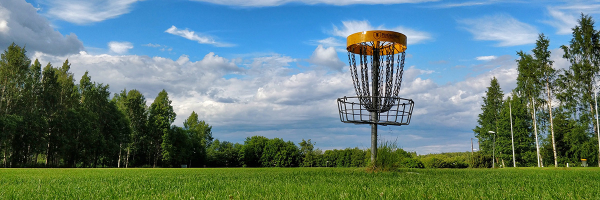 A disc golf basket is pictured on a grassy course surrounded by green leafy trees.