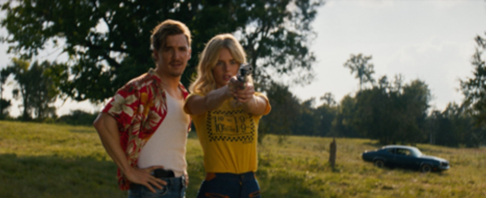 Film still from "Carolina Caroline." Woman is shown holding a gun as a man next to her looks on. A car appears in the background against a natural landscape.