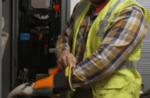 A lineworker in a plaid shirt and safety vest pulls on a pair of protective gloves.
