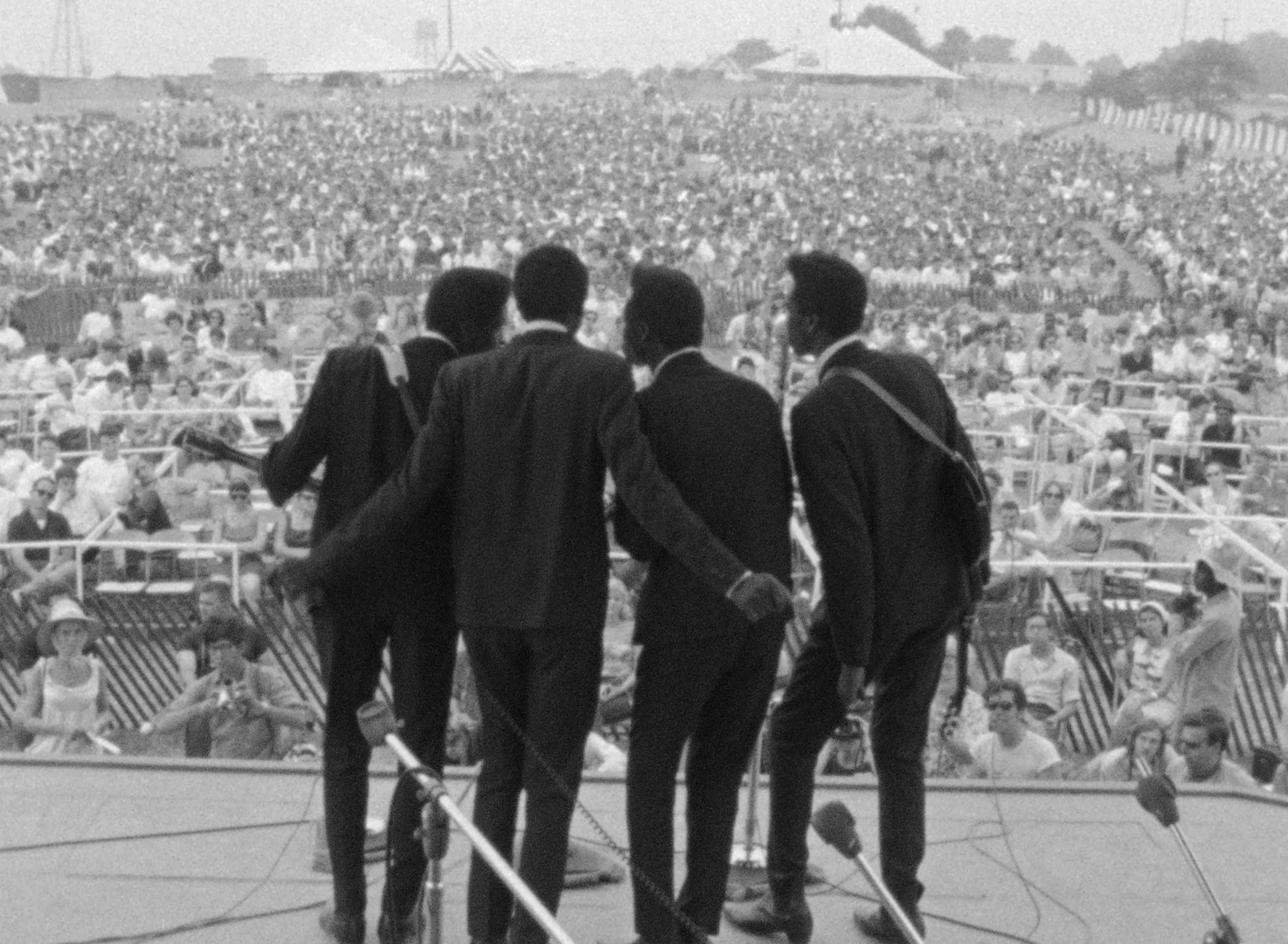 Image of four men photographed from the back singing into a single microphone to a sweeping crowd.