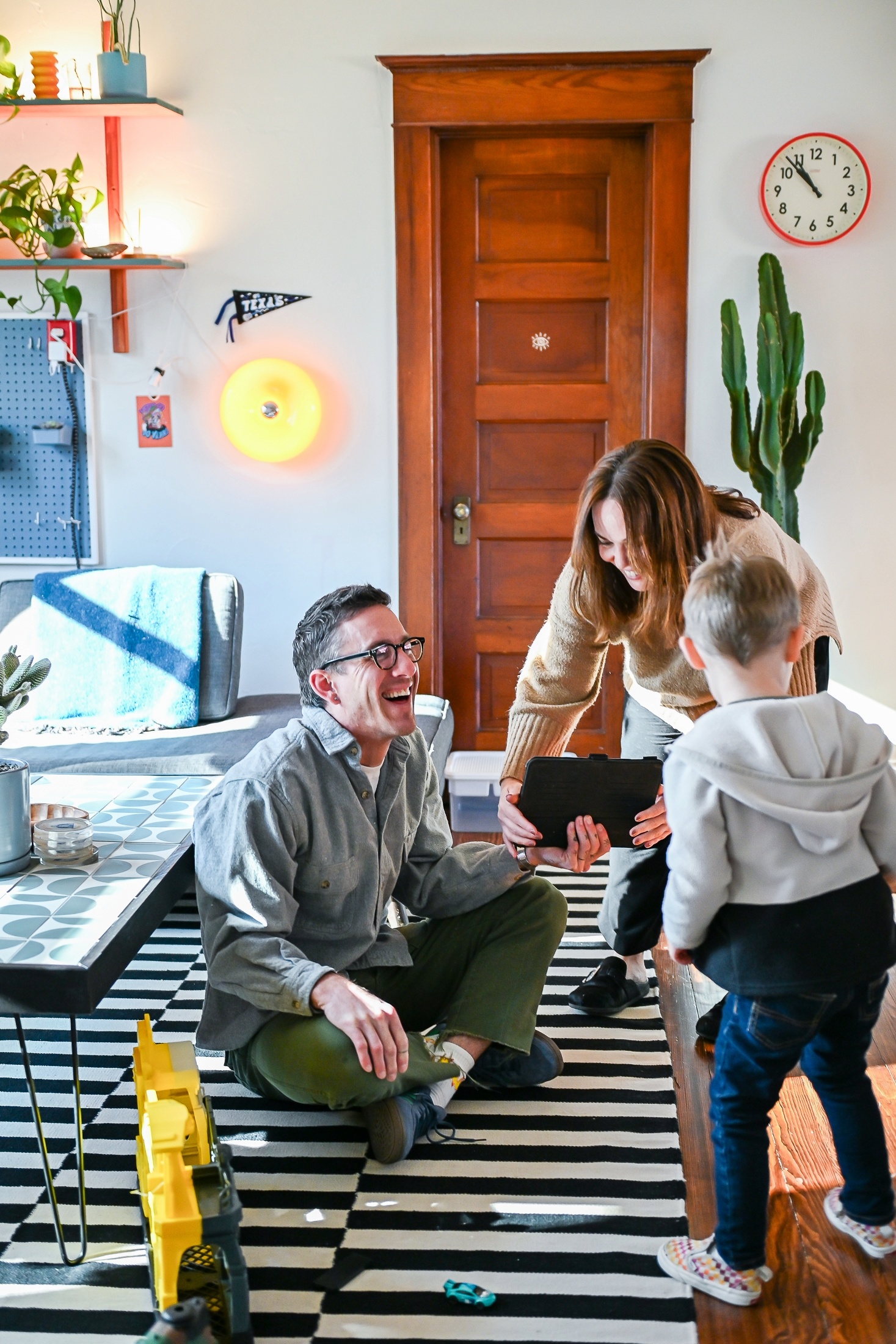 Laughing man is seated on the floor of his home as a smiling woman shows him a tablet and a small child approaches both of them.