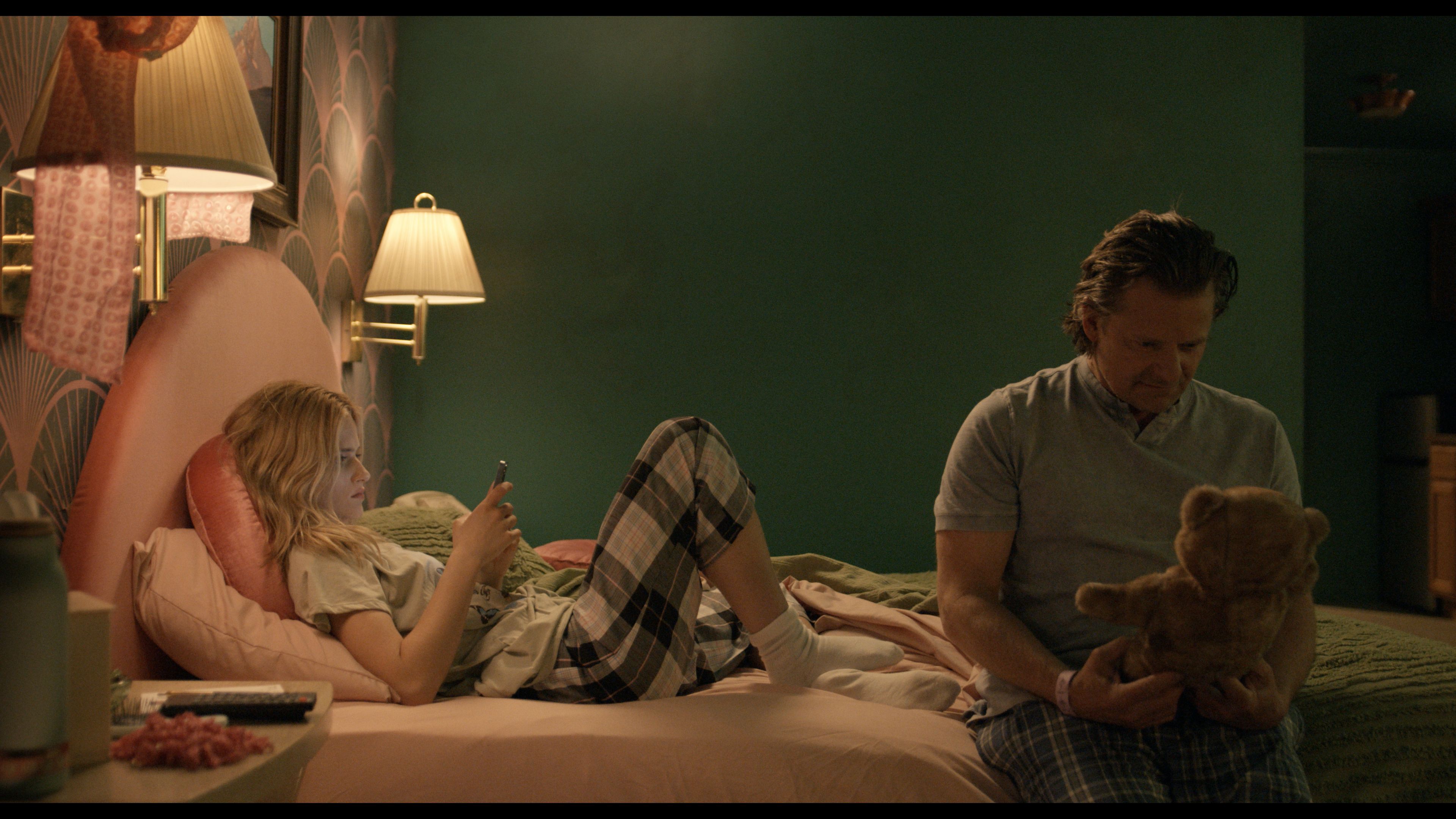 Film still from "She Dances" of a girl scrolling on her phone in her bedroom while her father sits on the end of the bed looking down at a teddy bear he is holding.