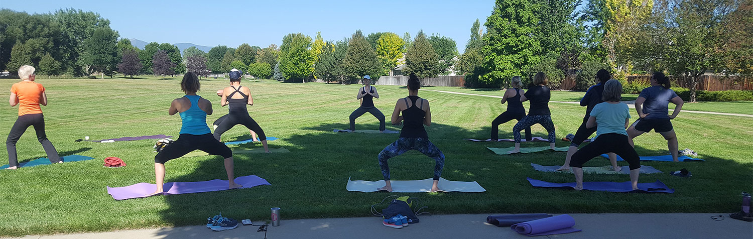 Group in park standing on mats in grass doing yoga poses with trees and mountain in background