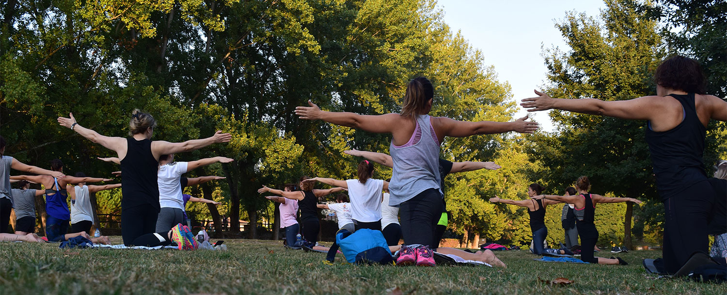 Group in park on knees in grass with arms stretched out during fitness class with trees in the background