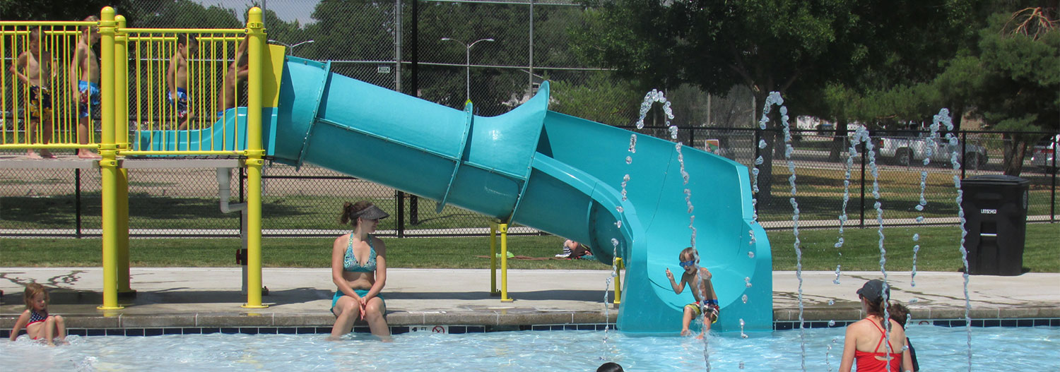 Bright aqua blue slide at outdoor Kanemoto pool with spray jets and people splashing