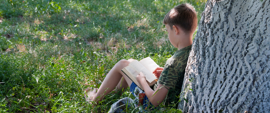 a Child reading a book and leaning against a tree in the grass.
