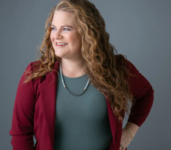 Midwestern comedian woman Sheila Chester with red long curly hair wearing a red jacket smiling.