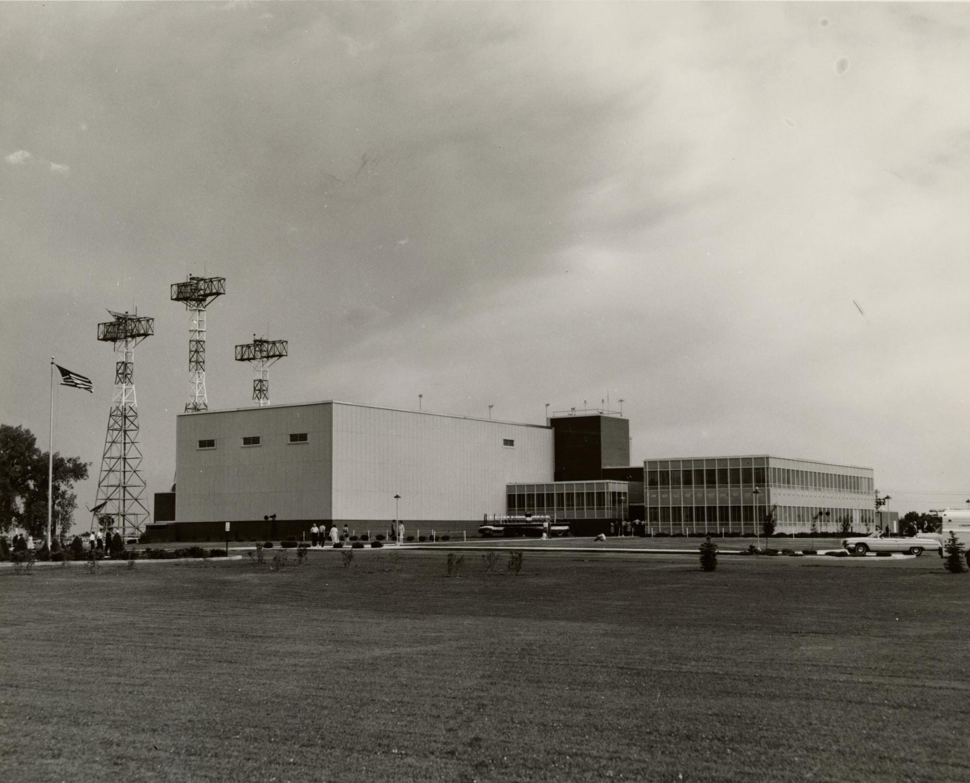 Black and White photograph of the exterior or the Denver Air Traffic Control Center in Longmont. Large radio towers seen on left, lawn in foreground, and large building with multiple wings at center.