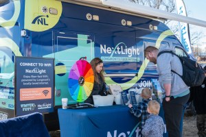 An adult with two small children visit NextLight's table at Winterfest, just outside the colorful NextLight van. A spin-the-wheel game is on the table. 