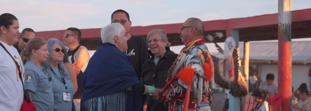 photo of seniors from Longmont shaking hands from a representative from the Wind River Reservation dressed in traditional Native American celebratory attire at an outside powwow in Wyoming
