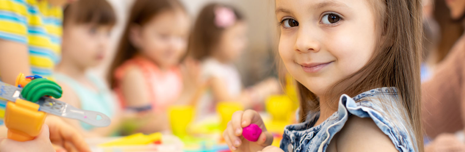 Happu child with brown eyes and brown hair holding a pink craft with other children doing play doh crafts