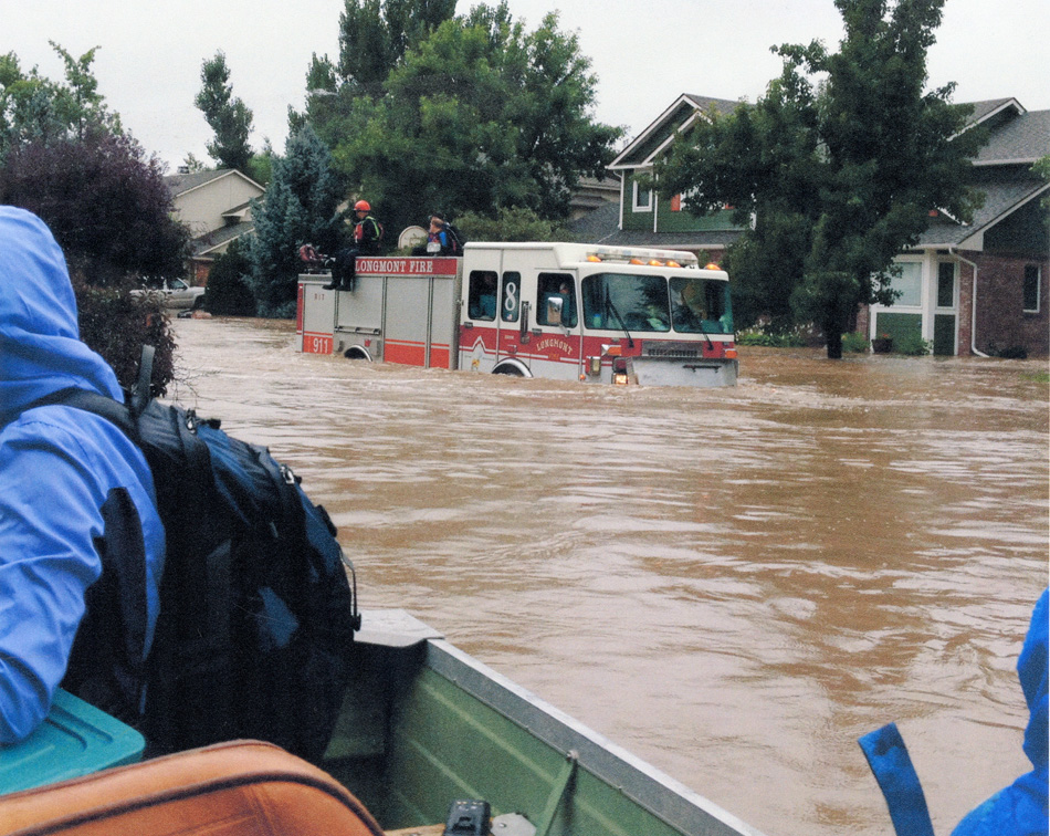 Color print of Longmont Fire Department fire engine with firefighters on top and driving through flooded streets of Longmont during flood of 2013. On right side of photo is a partial view of people in a boat.