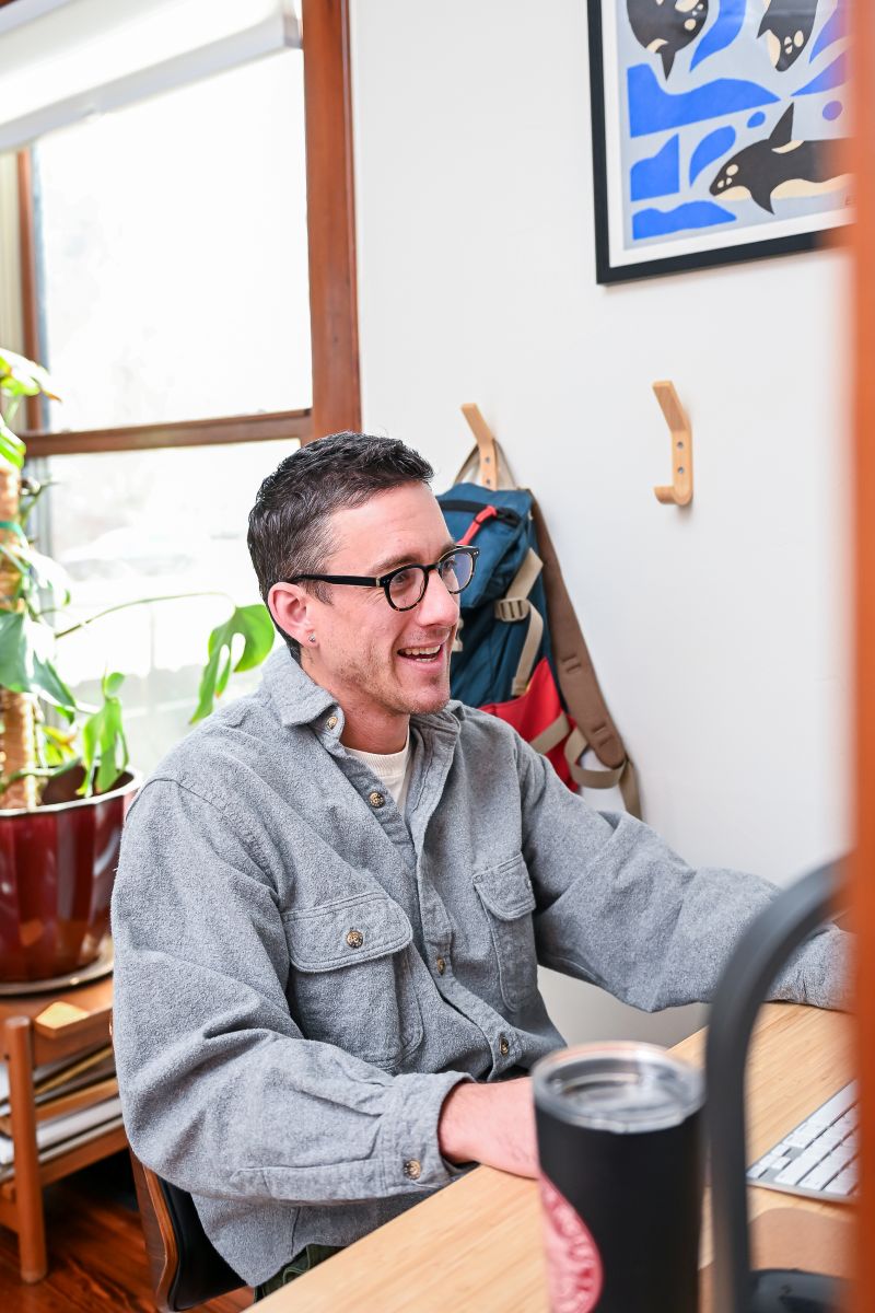 Smiling man sits at a computer with a sunny window behind him.