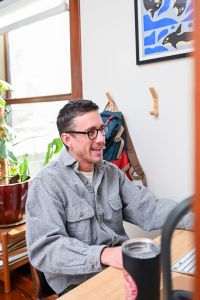 Smiling man sits at a computer with a sunny window behind him.