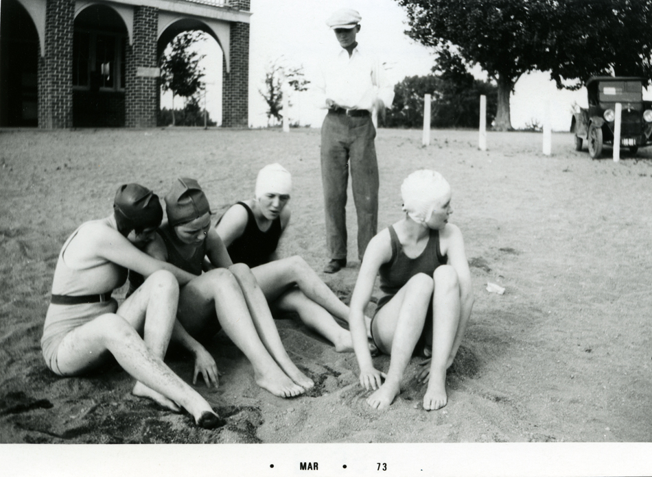 Four girls in swim suits and bathing caps sit on sand with a boy in long pants, white shirt, dark belt, and light colored cap standing behind them. In the background is a building with brick colonnades and trees and a dark car parked up against some white fence posts.