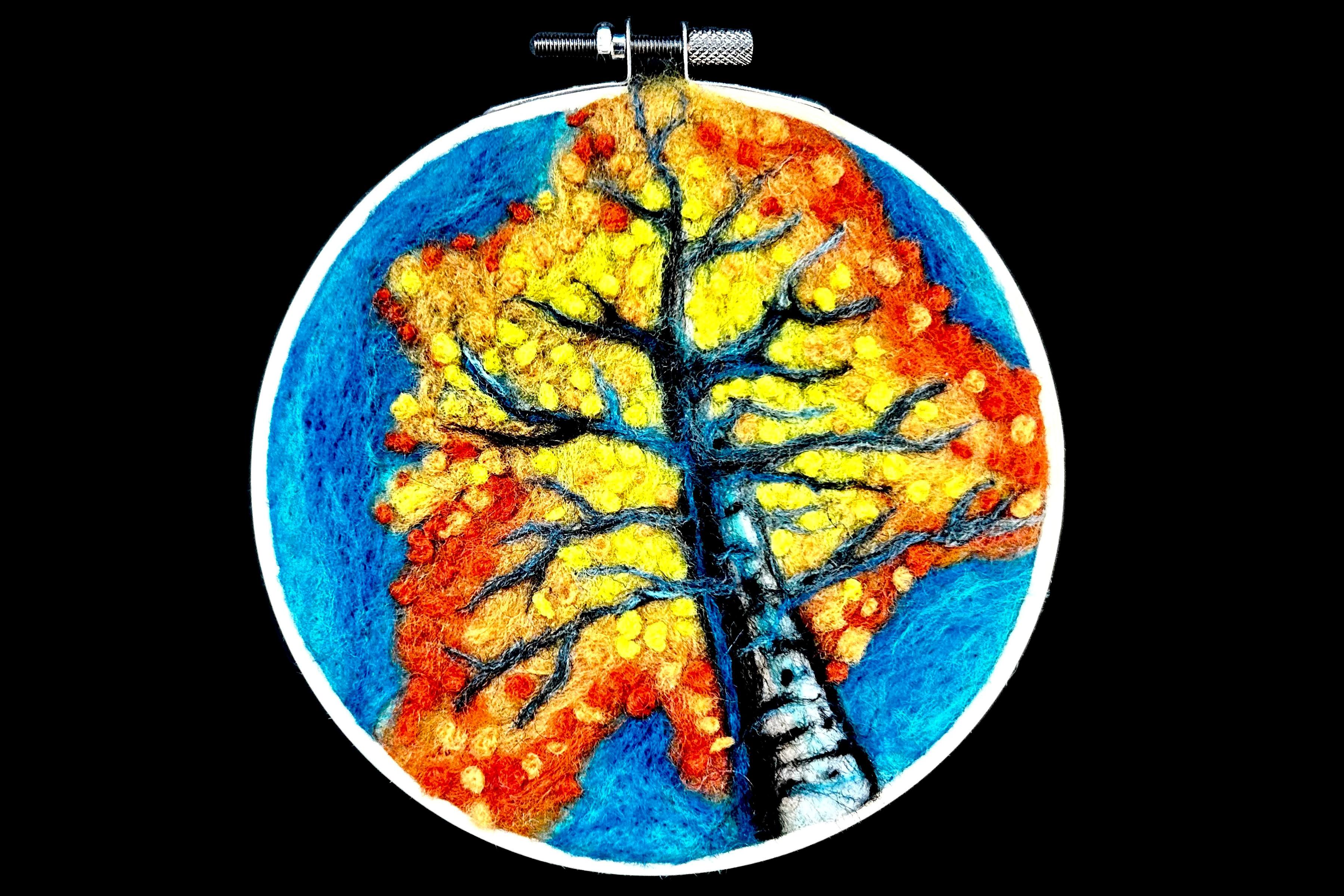 Image of a needlefelted autumn tree in an embroidery hoop rendered in black, white, yellow, orange, and blue felt.