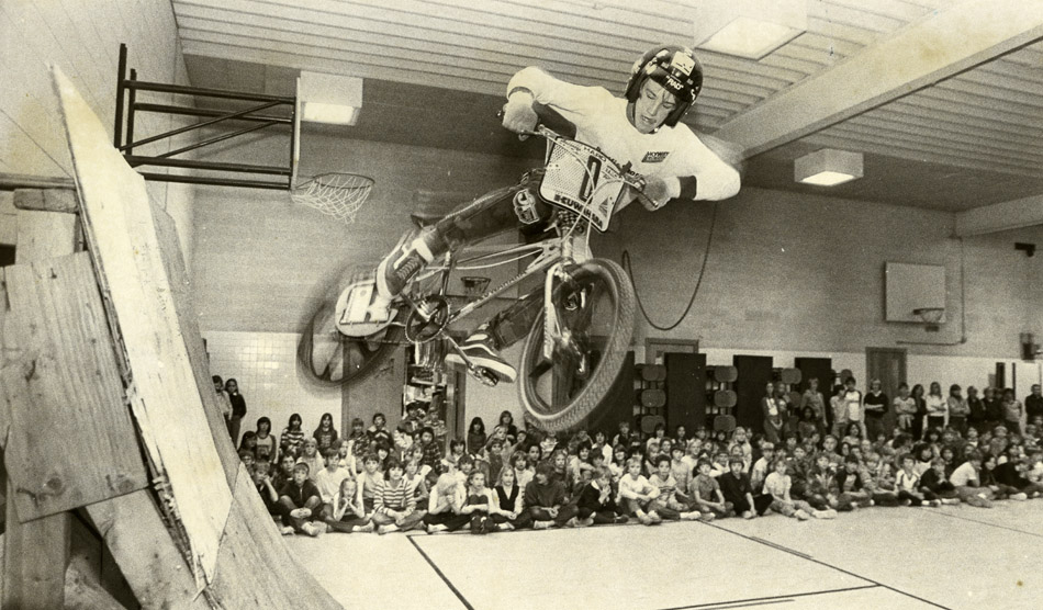 Black and white photo of a boy performing a bicycle trick in a school gym.