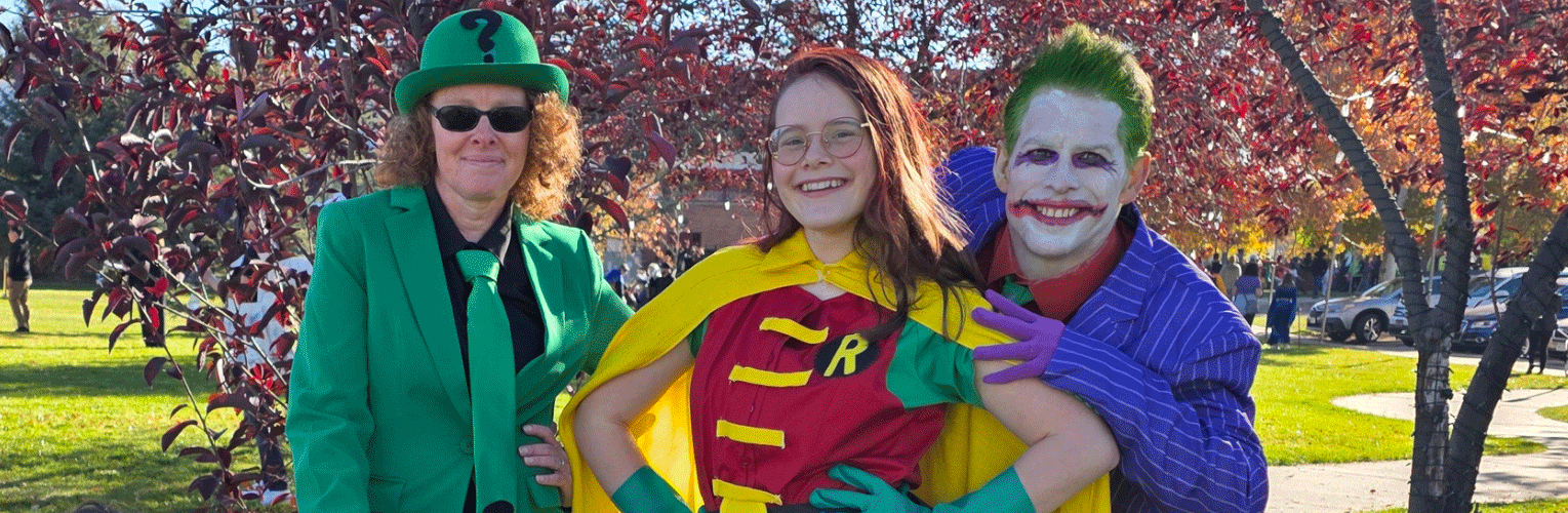 three adults in costume featuring the Riddler, Batgirl and the Joker in front of fall tree background