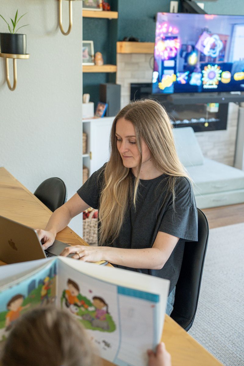 A woman works on her laptop at home while a young child reads a book across the table from her.