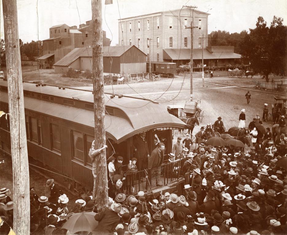 Teddy Roosevelt speaking from the back of a train in Longmont. There is a large crowd of people gathered around the platform at the back of the train, some with umbrellas. Roosevelt is bareheaded, and leaning forward over the railing slightly. In the background is the Farmer's Mill.