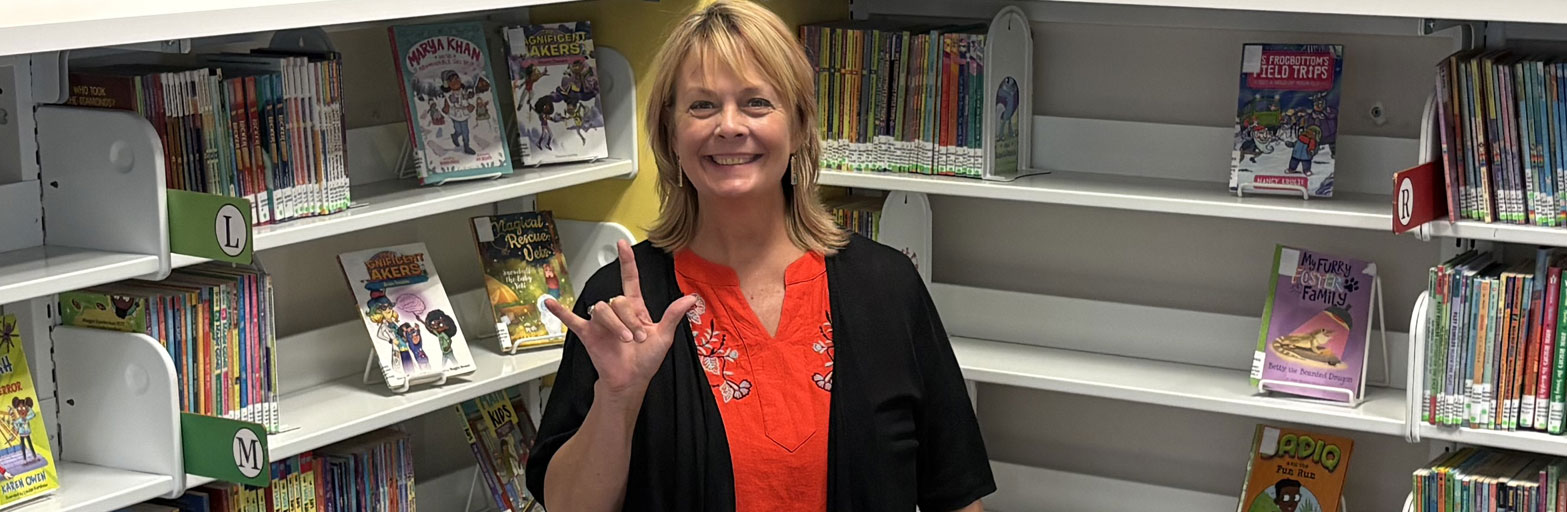 ASL instructor standing in front of a book shelf signing with her right hand.
