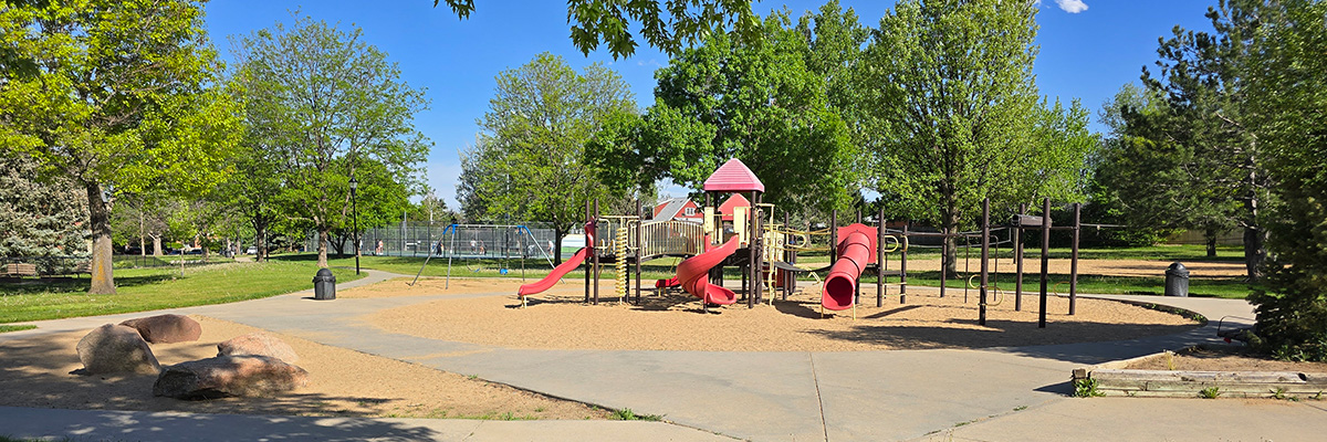 A colorful play structure, with bright red slides and roof, is seen in the middle of a wooded park.