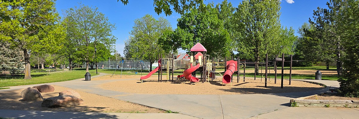 A colorful play structure, with bright red slides and roof, is seen in the middle of a wooded park.