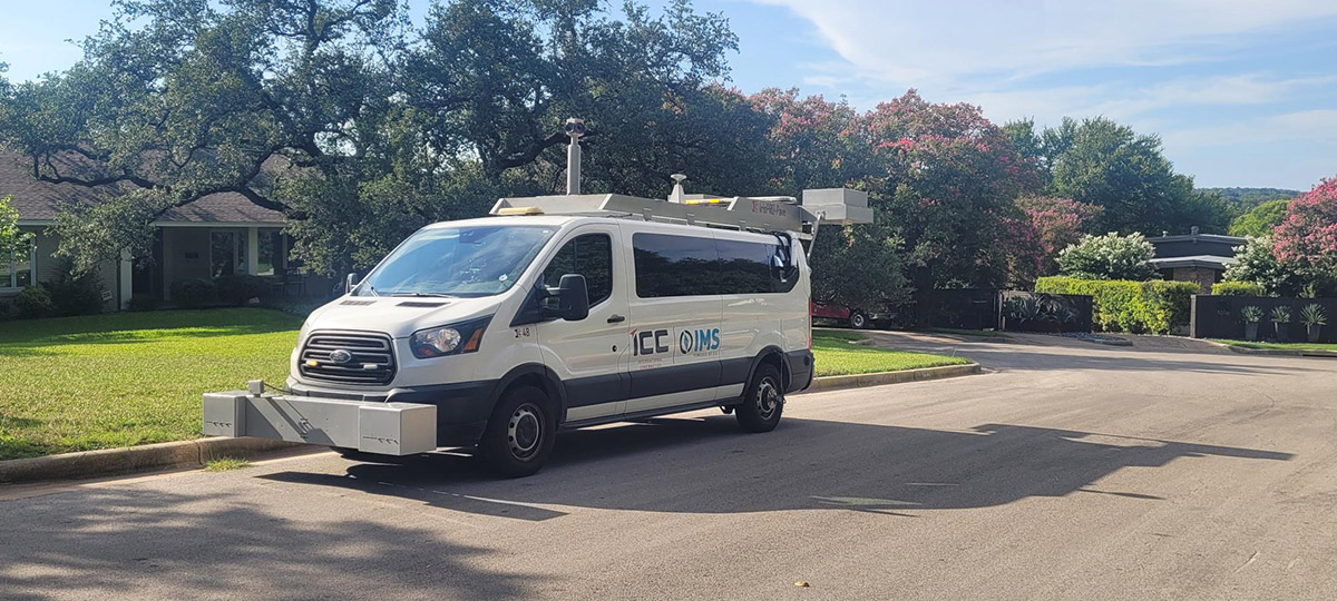 A white van equipped with cameras, sensors and other pavement assessment equipment is parked on a residential street lined with trees and homes. The specialized vehicle is used to scan and collect data about road conditions to help the City of Longmont evaluate pavement and plan future street maintenance.