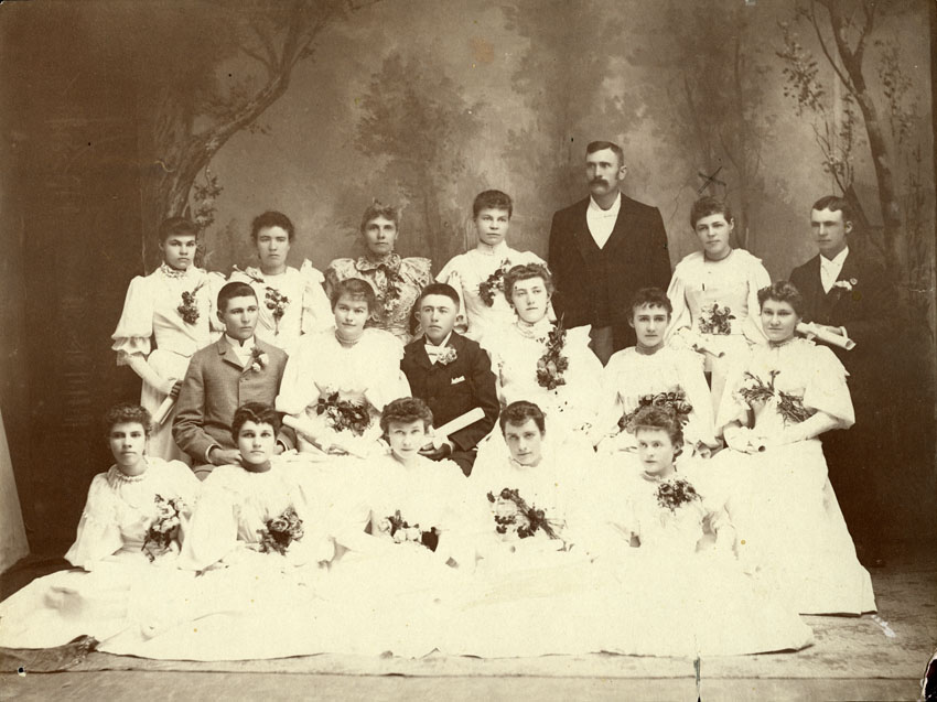 hotographic print of the Longmont High School graduating class from 1894. Photo is a studio print showing young women wearing long white dresses, each with flowers and diplomas. The young men are wearing suits and have a flower in their lapels.