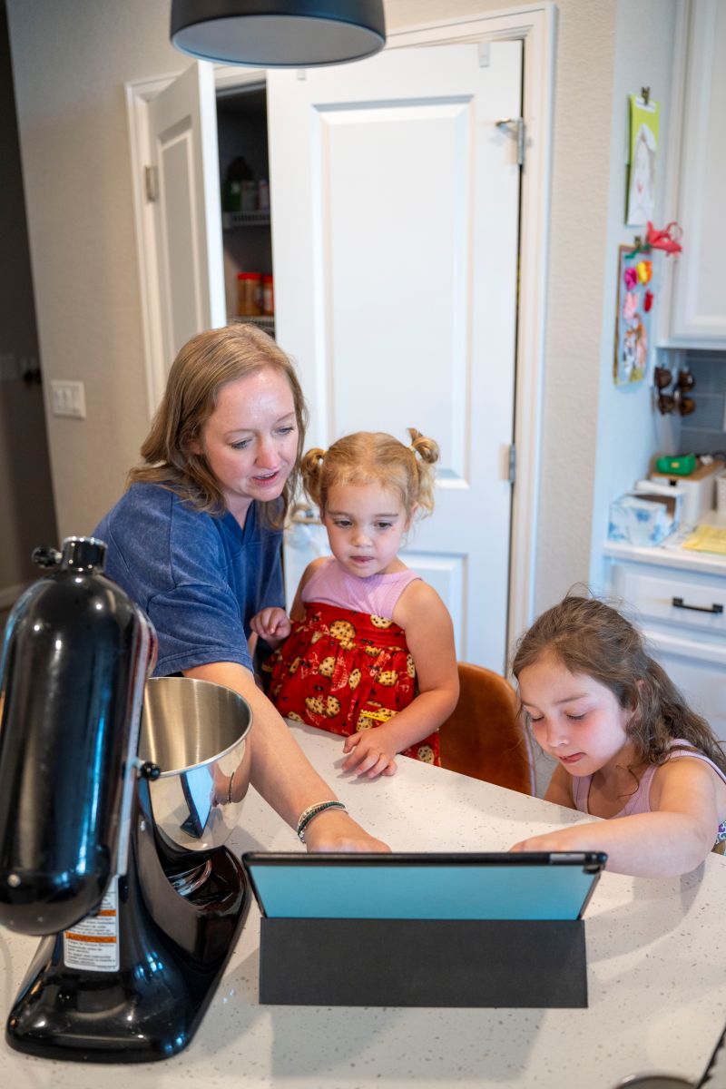 A woman and two young girls interact with a tablet on a kitchen counter