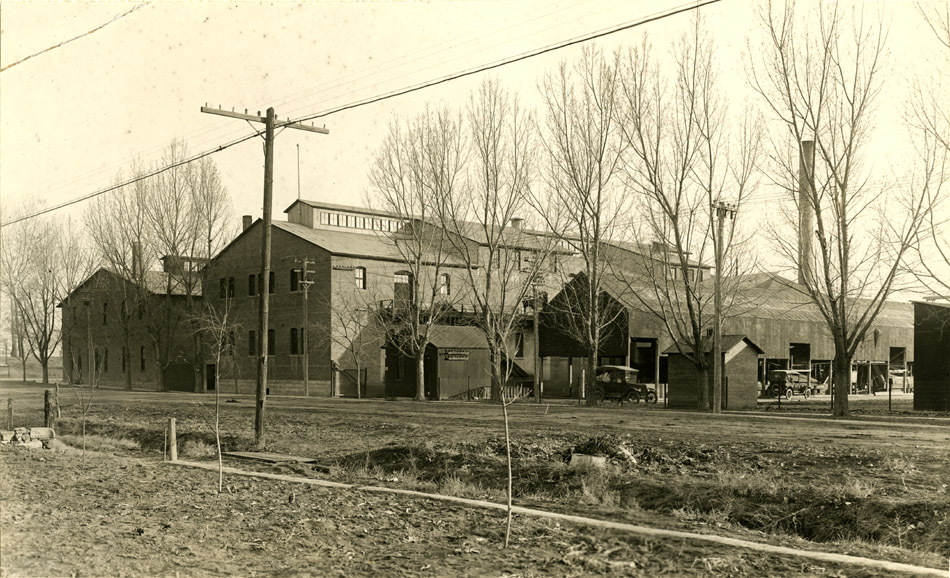 Print of the Empson Cannery. Photo shows main cannery building with clerestory windows along the roofline. Next to the main building are several sheds - some are small and enclosed, one is large and open-sided. Leafless trees are in front of the buildings.
